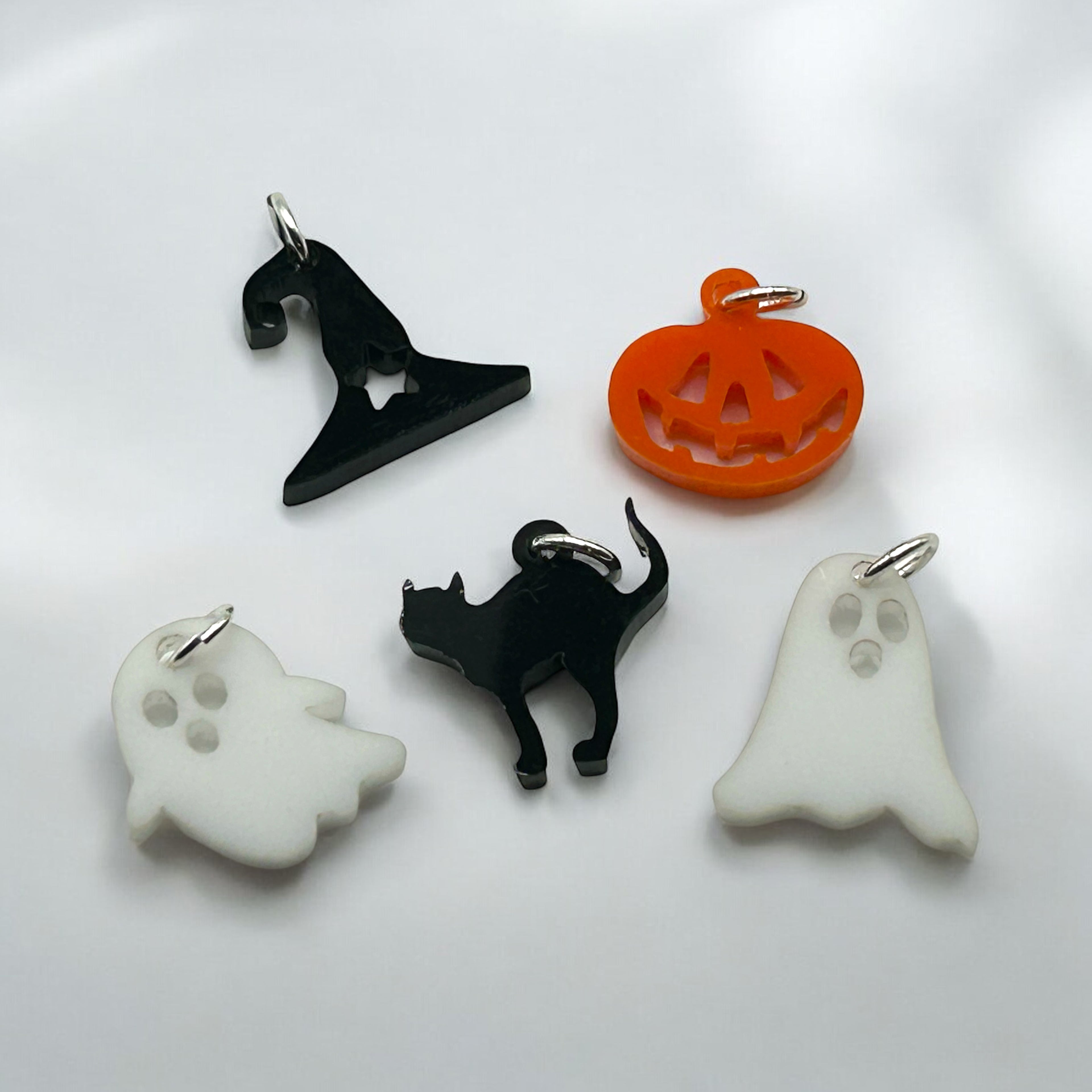 A collection of four acrylic charms with Halloween themes, including a pumpkin, a ghost, a witch's hat, and a cat, each with a sterling silver jump ring.