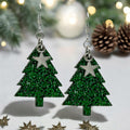 A pair of glittery green Christmas tree earrings with silver stars on top, displayed against a holiday-themed background with pine cones and lights.