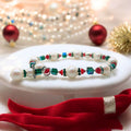 A festive Christmas-themed bracelet featuring a variety of colorful beads including pearls, red bicones, and emerald green crystals, displayed on a white stand with a red ribbon and Christmas decorations in the background.