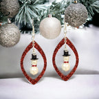 A photo of a Christmas-themed earring kit, featuring a red teardrop-shaped acrylic pendant with a white and black snowman figure inside, attached to a silver hook.