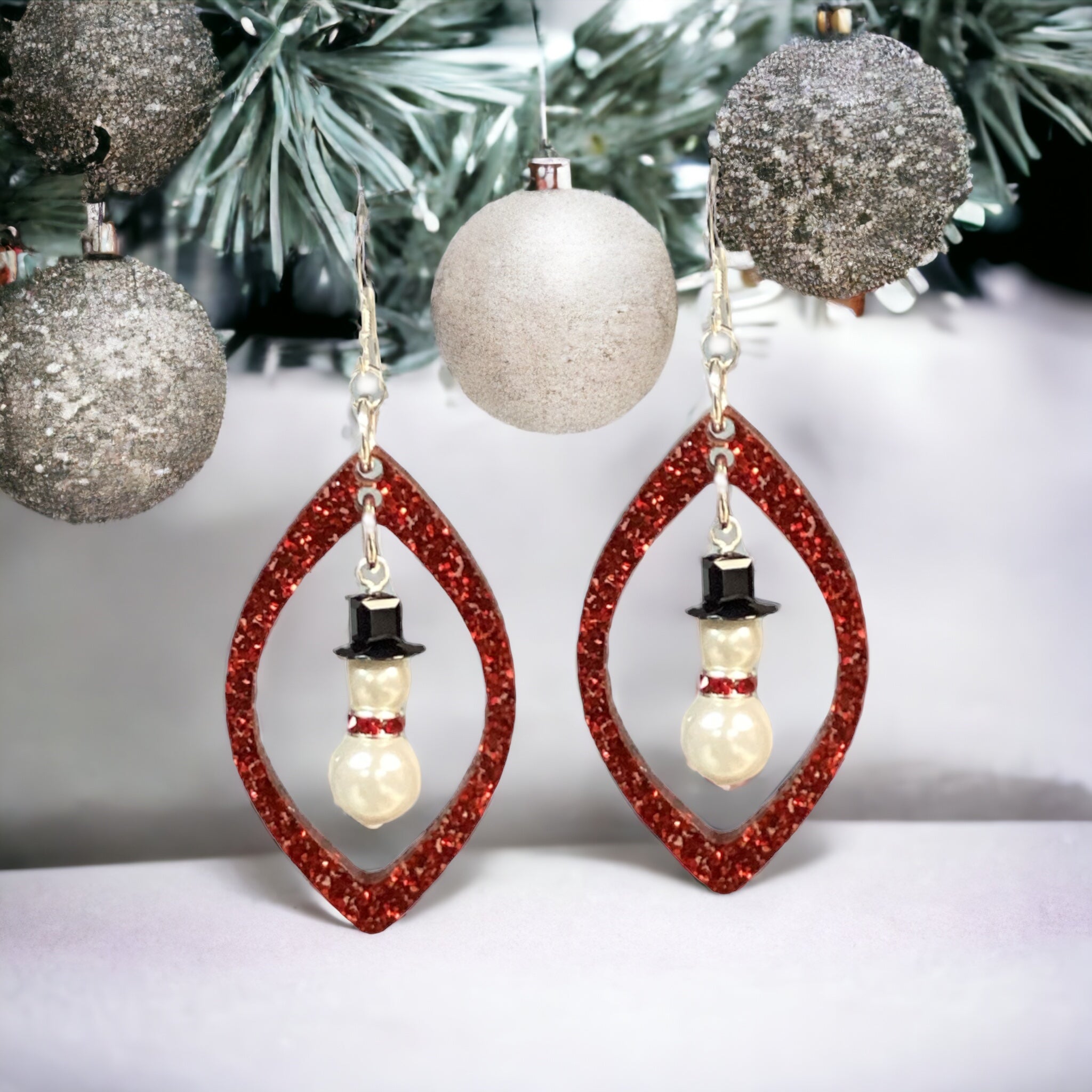 A photo of a Christmas-themed earring kit, featuring a red teardrop-shaped acrylic pendant with a white and black snowman figure inside, attached to a silver hook.