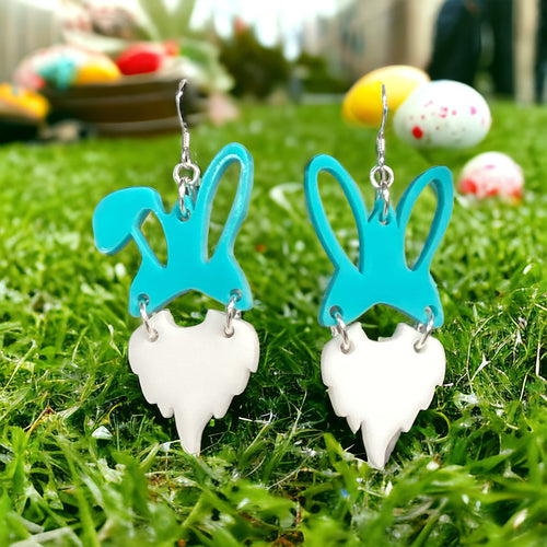 A pair of earrings crafted from acrylic gnomes in blue and white colors, displayed on green grass with Easter eggs in the background.