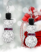 A photo showing a pair of pave snowman earrings with black, silver, and red beads against a festive Christmas themed background.